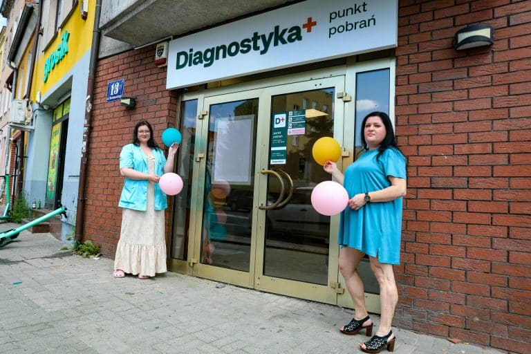 Dwie kobiety stoją przed wejściem do punktu pobrań Diagnostyka w Olsztynie, trzymając balony, na tle ceglanej ściany i szyby wejściowej.