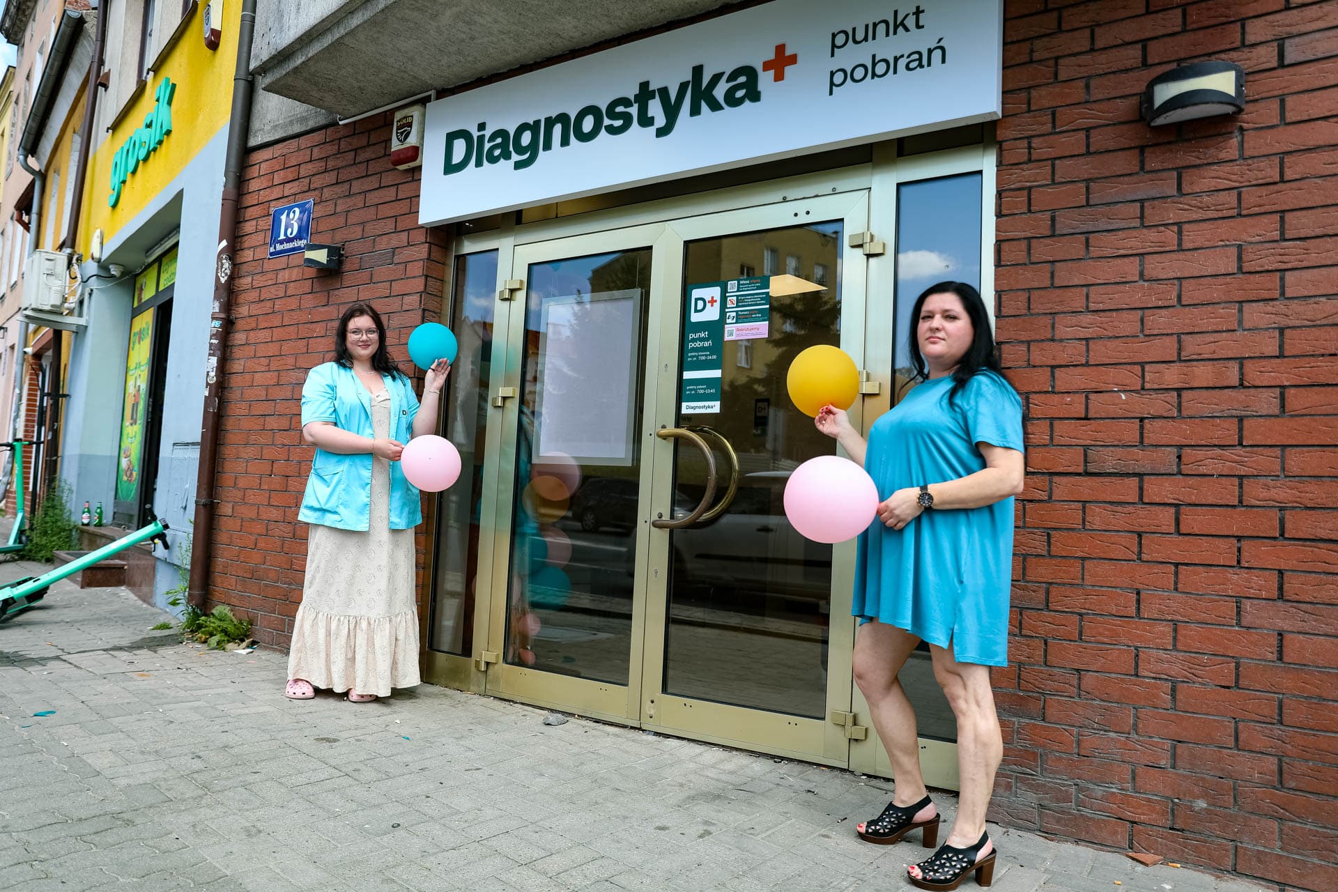 Dwie kobiety stoją przed wejściem do punktu pobrań Diagnostyka w Olsztynie, trzymając balony, na tle ceglanej ściany i szyby wejściowej.
