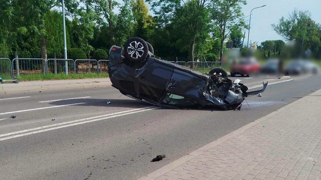 Rozbity czarny samochód leży na dachu na drodze, wokół są rozrzucone fragmenty pojazdu.