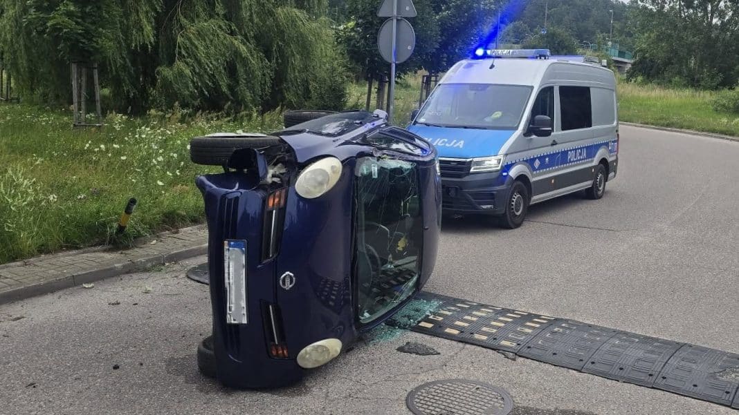 Rozbity samochód leży na boku na ulicy, obok policyjny radiowóz z włączonymi światłami.