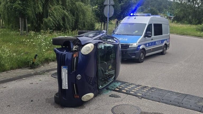 Rozbity samochód leży na boku na ulicy, obok policyjny radiowóz z włączonymi światłami.