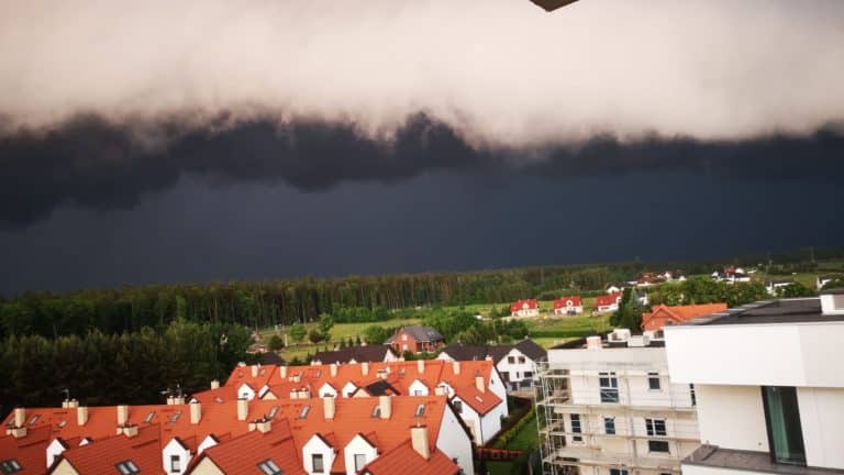 Ciemne, burzowe chmury nad miastem z czerwonymi dachami i zielonymi terenami w tle.