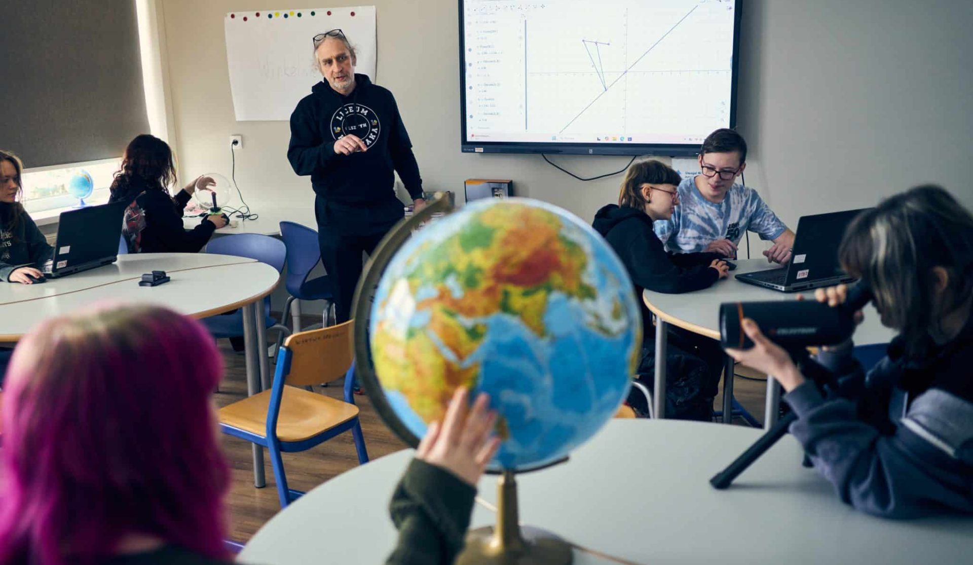 Nauczyciel prowadzi zajęcia z uczniami w klasie, na ekranie wyświetlane są wykresy, w tle globus na stole.
