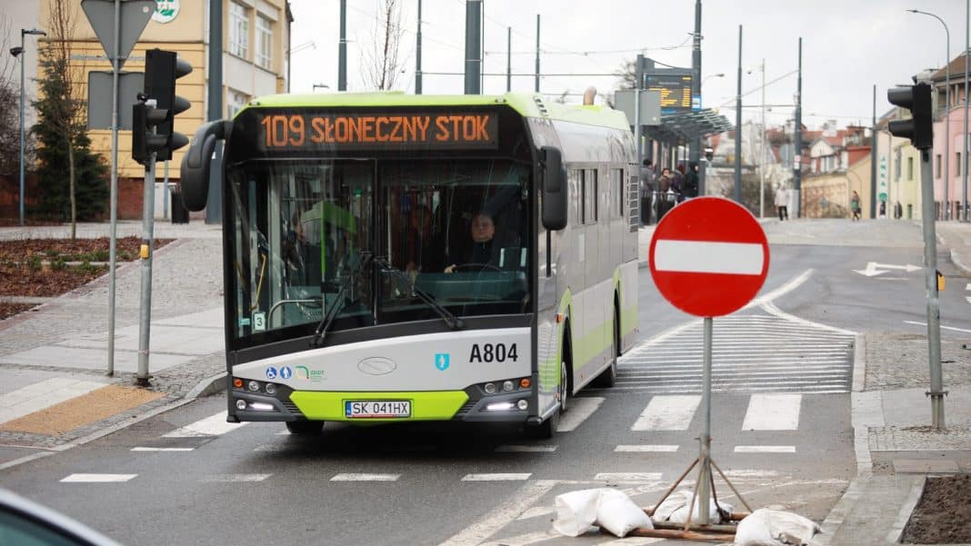 Autobus miejski na przystanku w mieście, w tle budynki i ludzie czekający na autobus.
