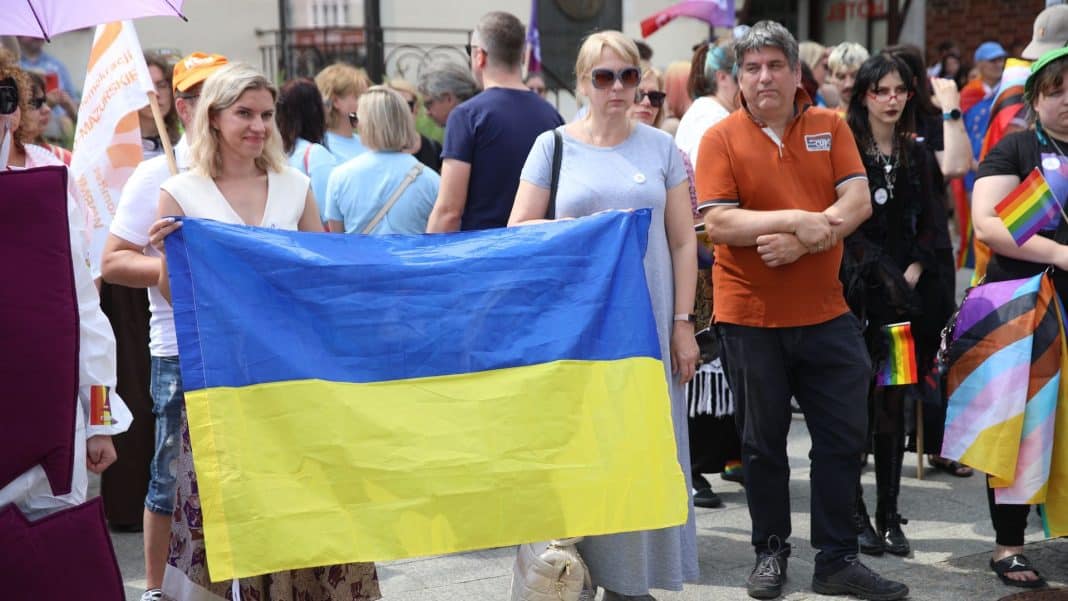 Grupa ludzi na zgromadzeniu w centrum Olsztyna, niektórzy trzymają flagę Ukrainy, w tle tłum uczestników.
