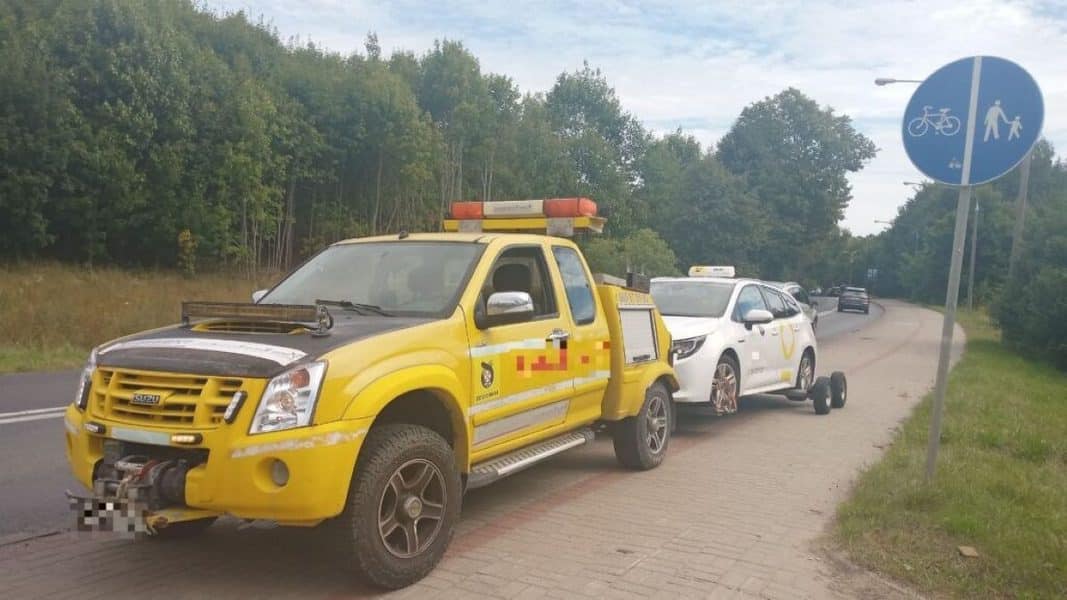 Żółta taksówka zaparkowana na drodze dla rowerów, odholowana na lawetę, w tle las i znak rowerowy.