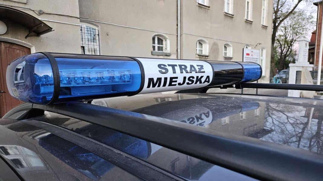 Na zdjęciu widoczny jest policyjny samochód z niebieskim światłem na dachu, na tle budynków mieszkalnych.