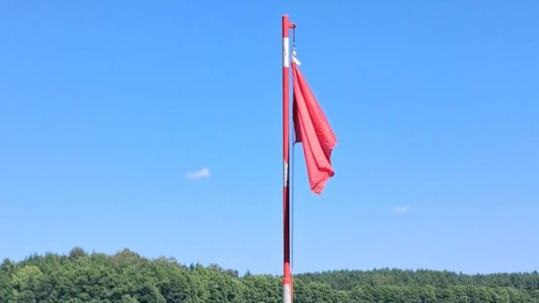 Flaga na maszcie na tle nieba i zielonych drzew, bez ludzi na zdjęciu.