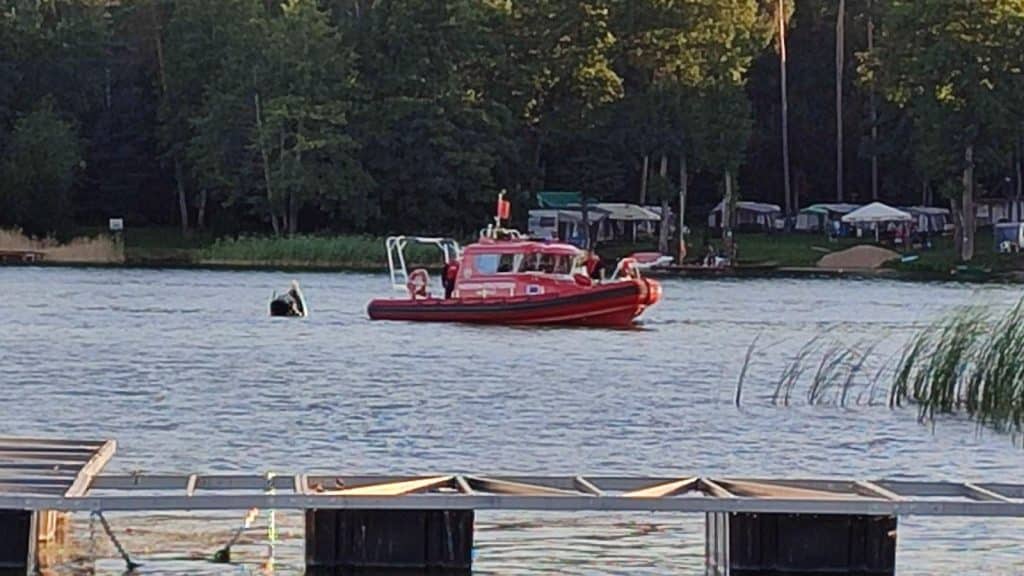 Czerwony ponton ratunkowy na jeziorze z widokiem na plażę i las w tle.