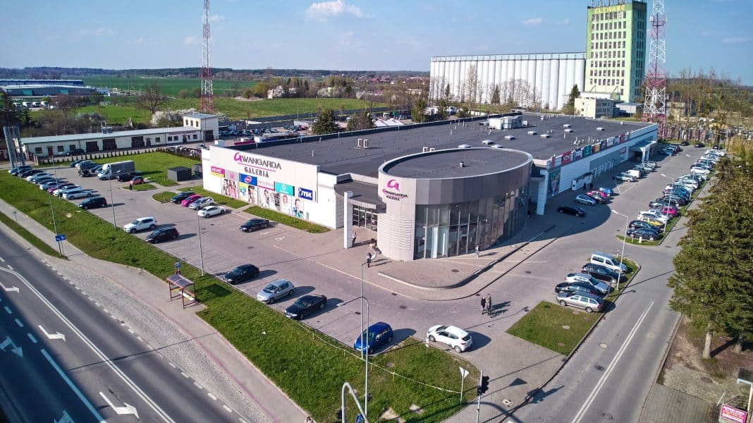 Aerial view of a large shopping mall with parking lot, surrounded by greenery and open fields.