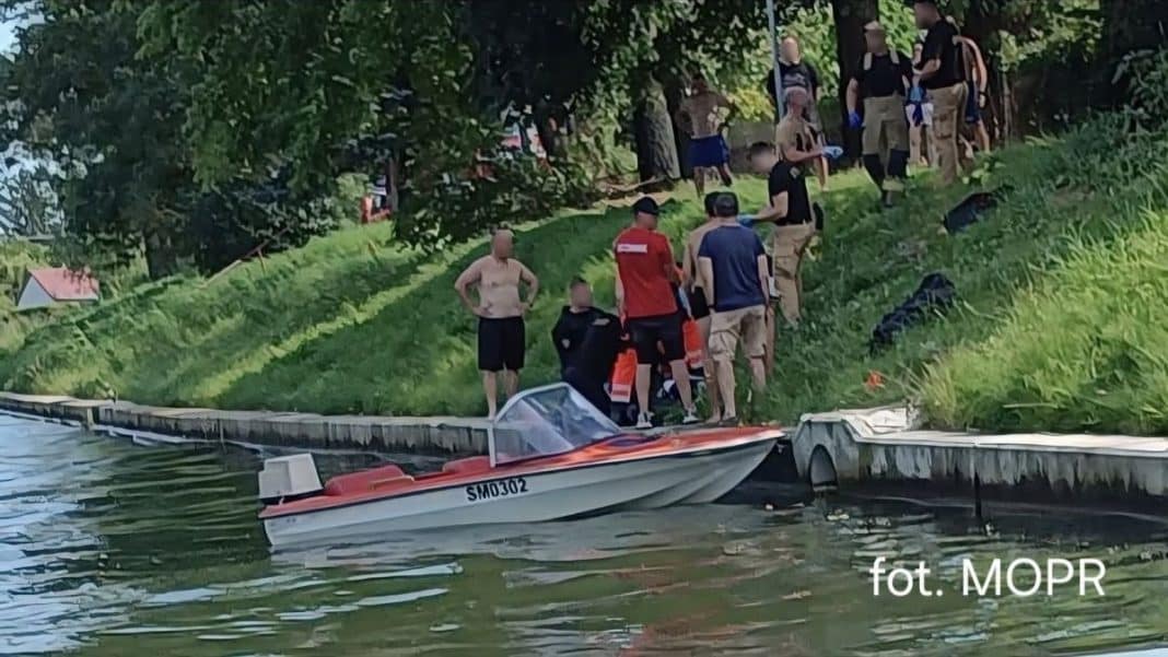 Grupa świadków i ratowników na brzegu kanału, z łodzią ratunkową w tle, podczas akcji ratunkowej w Giżycku.