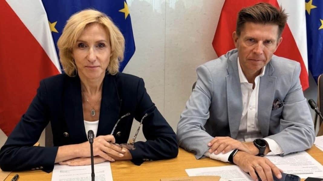 Dwie osoby siedzą przy stole z dokumentami, za nimi flagi Polski i Unii Europejskiej.
