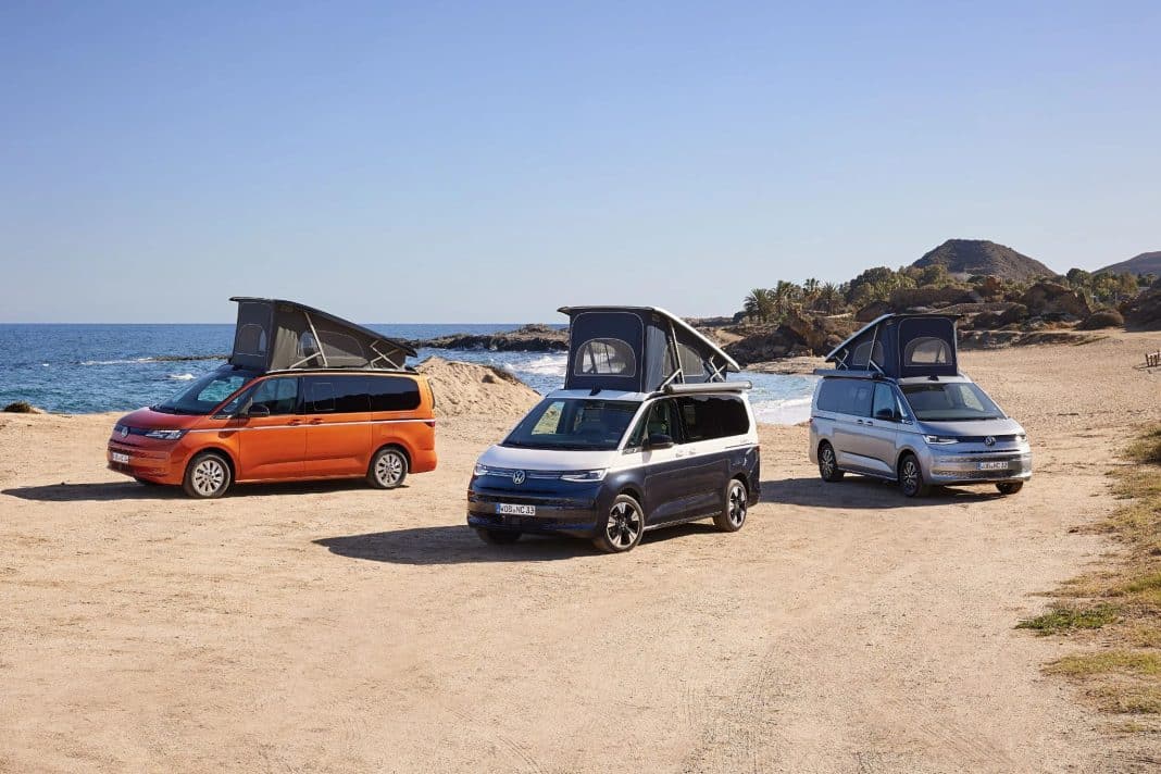 Trzy samochody typu camper na plaży nad jeziorem, z namiotami na dachach, w tle skały i roślinność.