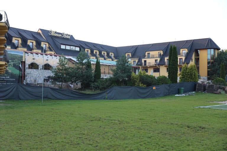 Widok na hotel Manor w Olsztynie z ogrodzonym terenem i drzewami, uszkodzona ściana budynku podczas prac budowlanych.