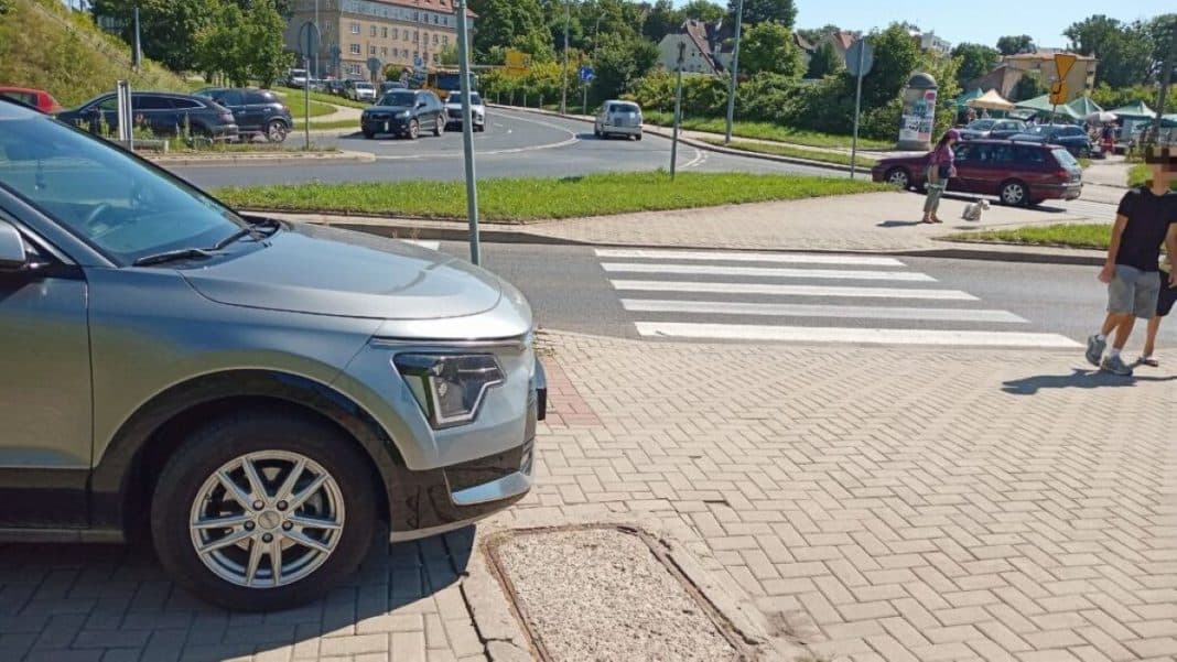 Samochód zaparkowany na chodniku przy targowisku, w tle ludzie i ruch uliczny.