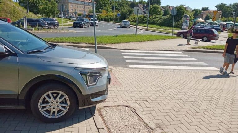 Samochód zaparkowany na chodniku przy targowisku, w tle ludzie i ruch uliczny.