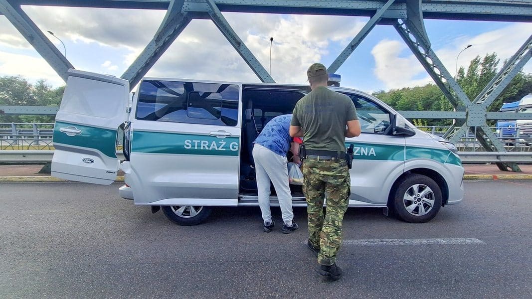 Policjant zatrzymuje mężczyznę przy samochodzie na drodze, w tle most i niebo z chmurami.