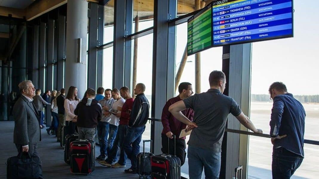 Pasażerowie czekający na lotnisku przy tablicy odlotów, w tle okno z widokiem na pas startowy.