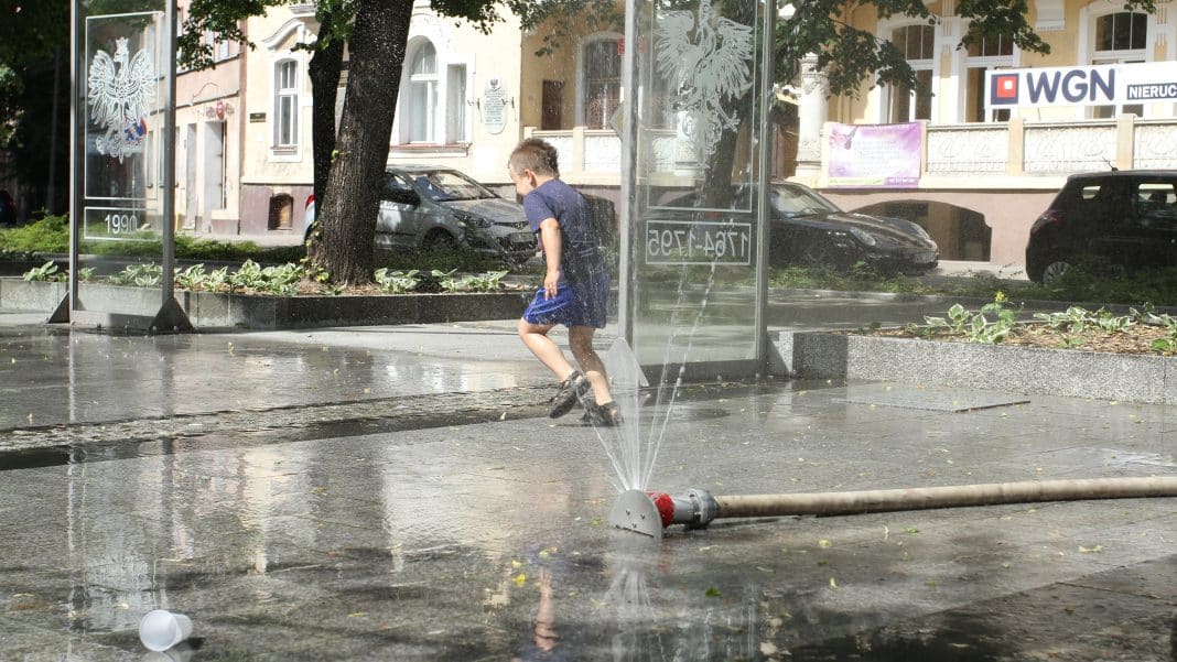 Dziecko bawi się pod wodnym prysznicem na ulicy w upalny dzień, w tle samochody i budynki.
