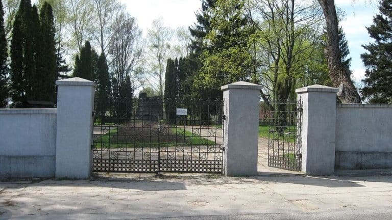 Widok na ogrodzenie i wejście na cmentarz wojenny w Olsztynie, otoczone drzewa i zielenią.