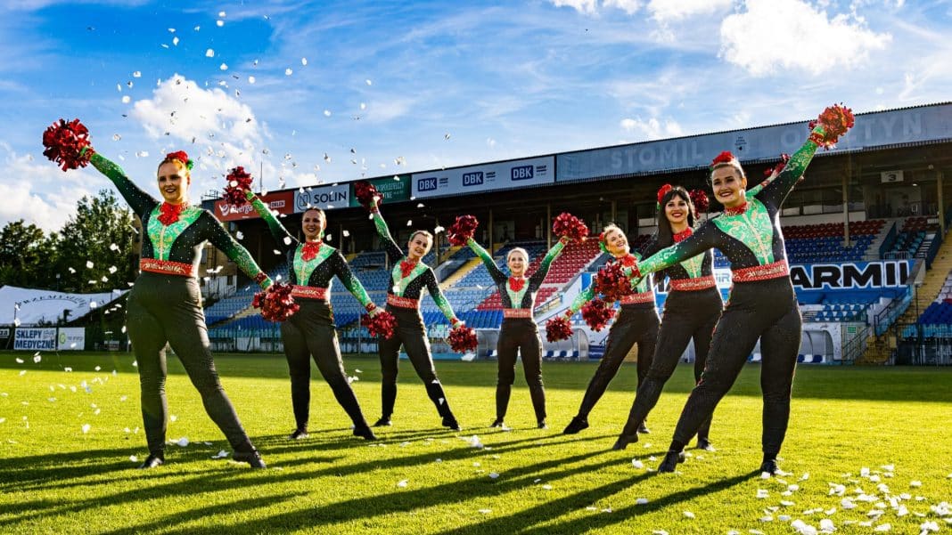 Grupa cheerleaderek w strojach w stylu świątecznym na boisku, rzucają confetti podczas wydarzenia sportowego.