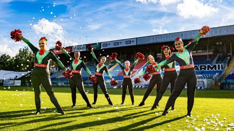 Grupa cheerleaderek w strojach w stylu świątecznym na boisku, rzucają confetti podczas wydarzenia sportowego.