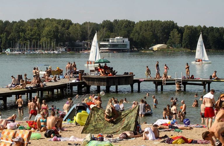 Tłum ludzi na plaży i na pomoście nad jeziorem Ukiel, w tle żaglówki i budynki.