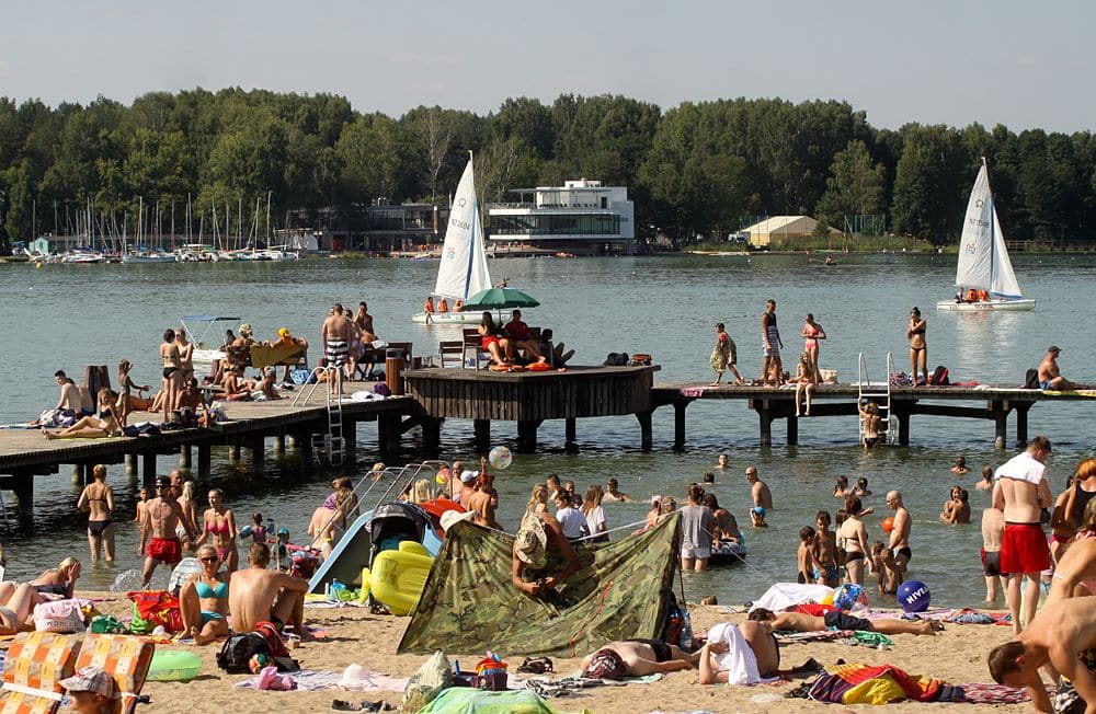 Tłum ludzi na plaży i na pomoście nad jeziorem Ukiel, w tle żaglówki i budynki.