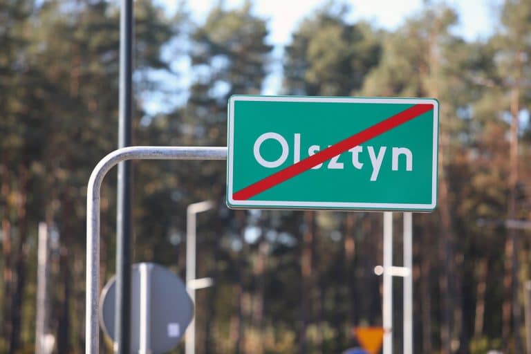 Znak końca miasta Olsztyn na drodze, otoczony lasem i drzewami w tle.