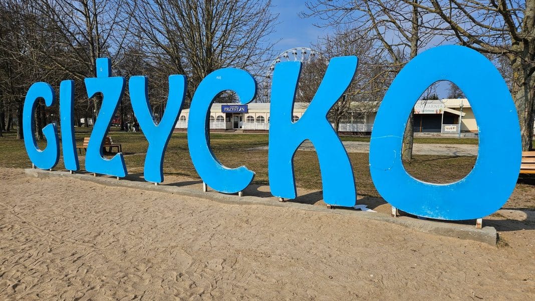 Duży, niebieski napis 'Giżycko' na tle parkowego otoczenia, bez ludzi, w słoneczny dzień.