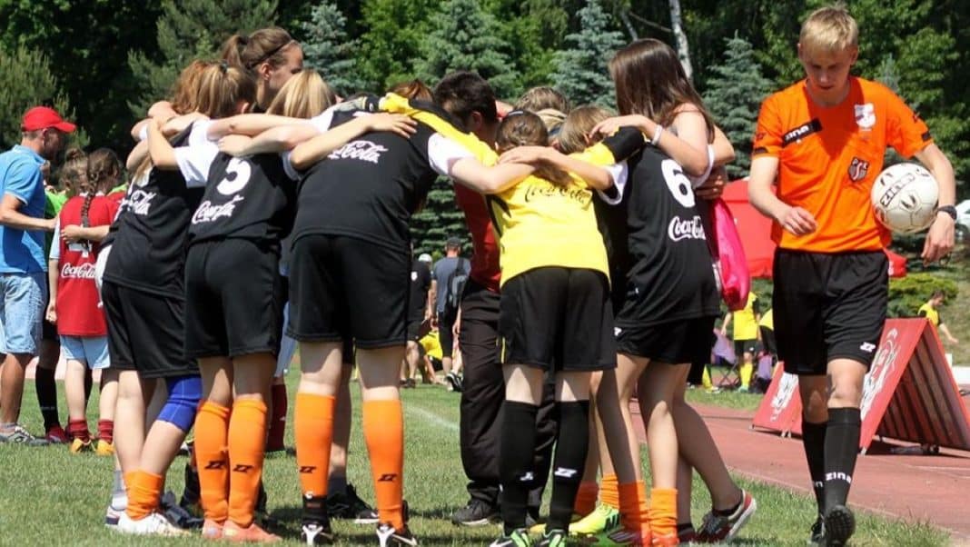 Grupa młodych sportowców w kółku na boisku, w tle drzewa i inni uczestnicy wydarzenia.