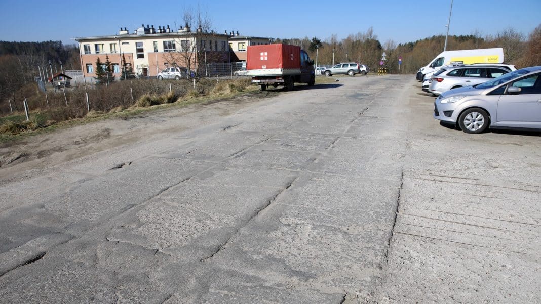 Pusty parking z nierówną, pękniętą nawierzchnią, w tle budynek i samochody na parkingu.