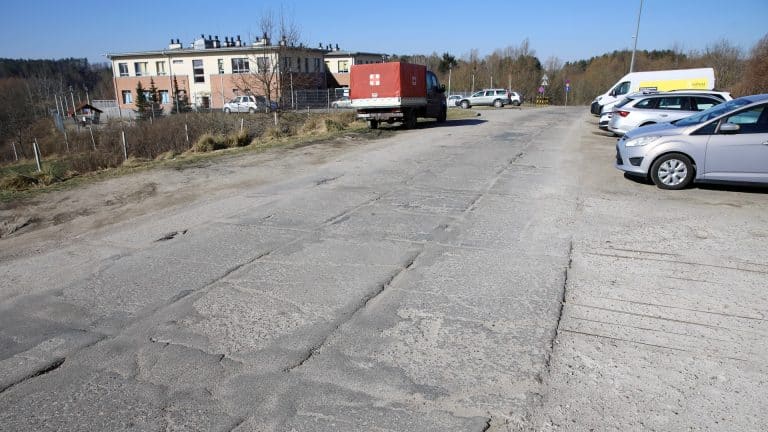 Pusty parking z nierówną, pękniętą nawierzchnią, w tle budynek i samochody na parkingu.
