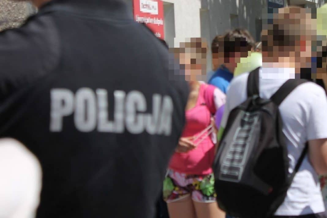 Grupa dzieci na osiedlu w Olsztynie, z widocznym policjantem w tle, na tle bloków mieszkalnych.