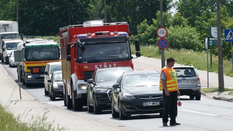 Kierowcy czekają w korku na drodze, obok stoi strażak w odblaskowej kamizelce.