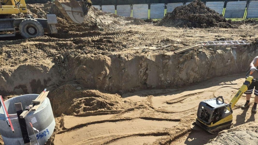 Prace budowlane na placu z ciężkim sprzętem i wykopami, w tle ogrodzenie i nowoczesny plac budowy.