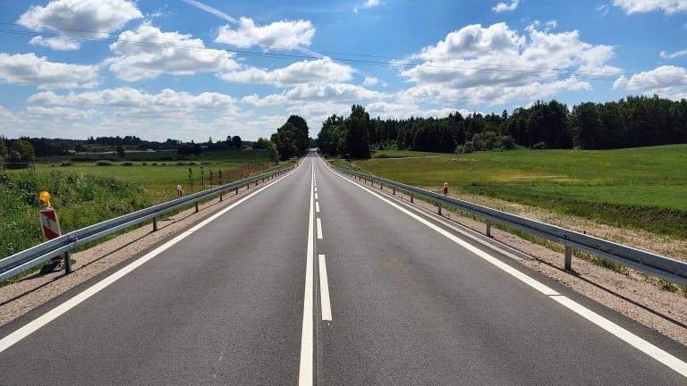 Długa, prosta droga krajowa w otoczeniu zielonych pól i drzew pod błękitnym niebem z chmurami.