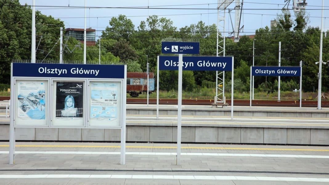 Peron na peronie dworca kolejowego z tablicami informacyjnymi i oznaczeniem Olsztyn Główny.