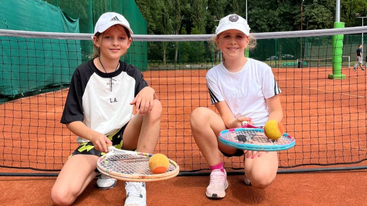 Dwie młode tenisistki na korcie trzymające rakiety i piłki, uśmiechnięte, w sportowych strojach i czapkach.