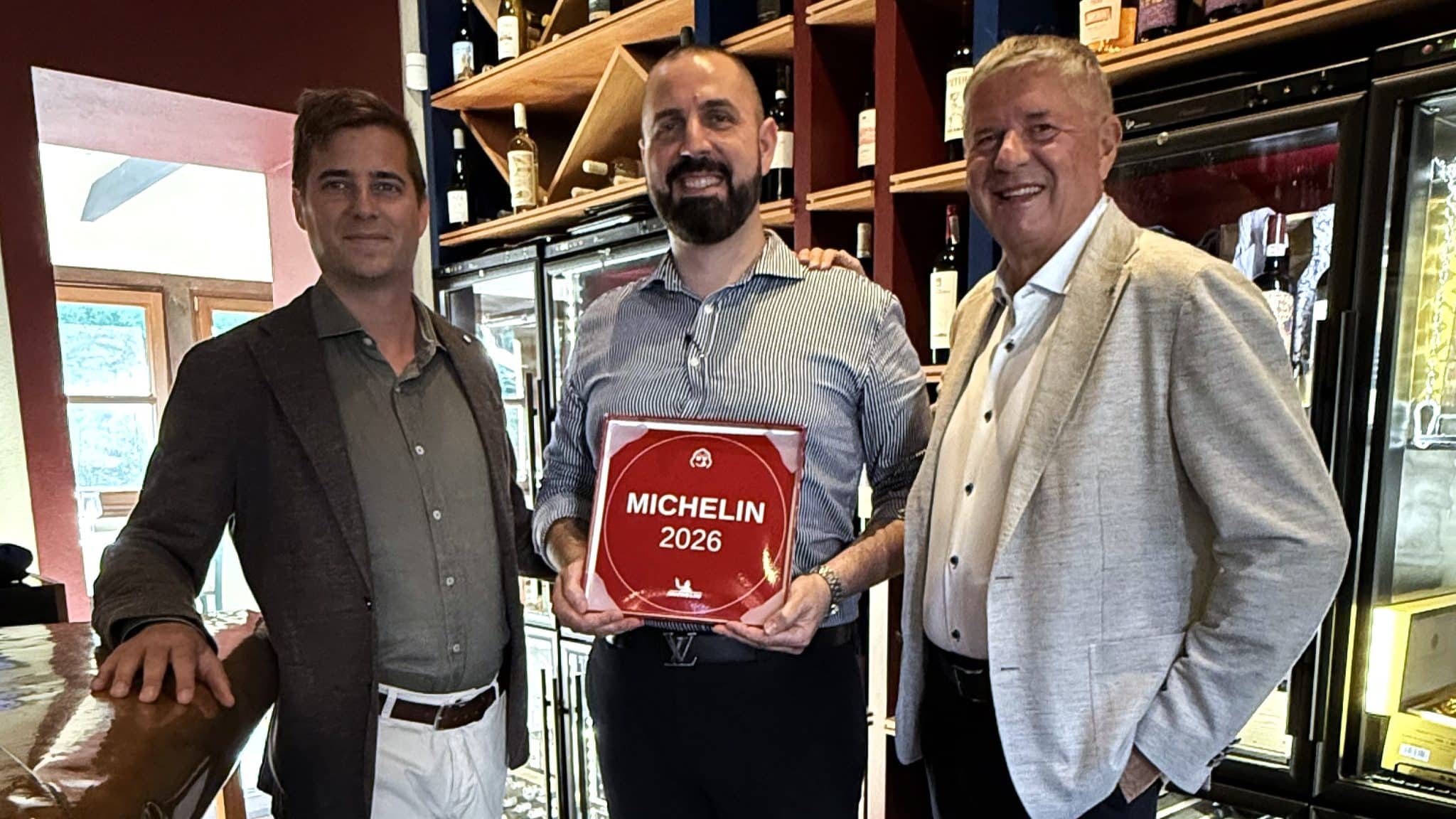 Trzej mężczyźni stojący w restauracji, jeden z nich trzyma tabliczkę z napisem 'Michelin 2026'.
