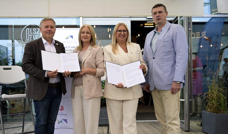 Grupa czterech osób trzymających dokumenty podczas wydarzenia na zewnątrz, w tle logo i wejście do budynku.