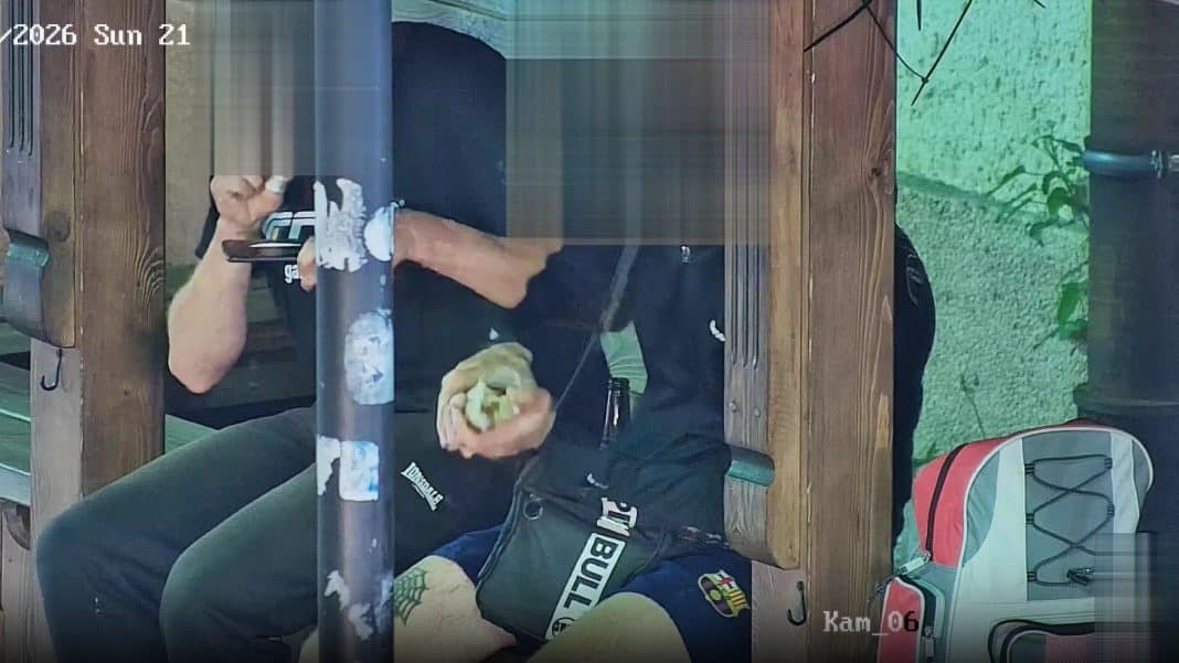 Mężczyzna siedzący przy stoliku, trzymający telefon i jedzenie, w tle drewniany stół i torba na zewnątrz.