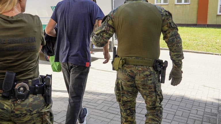 Funkcjonariusze Straży Granicznej odprowadzają cudzoziemca, w tle budynek mieszkalny.