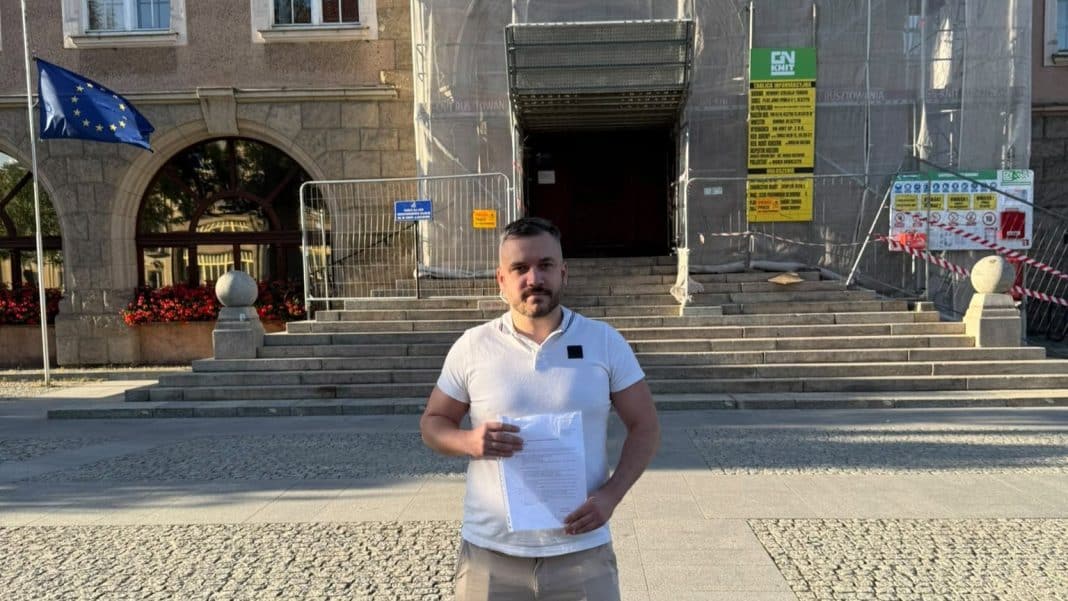 Mężczyzna trzymający dokumenty na tle budynku z wejściem i schodami, wokół ogrodzenie i informacje o pracach remontowych.
