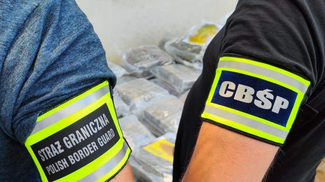 Ujęcie służb granicznych i policji przy zabezpieczonym magazynie narkotyków, w tle widoczne pakiety substancji.