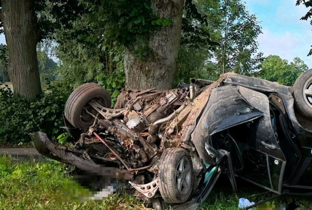 Rozbity samochód BMW po wypadku, leży na boku przy drzewie, uszkodzony i porozrzucany na poboczu.