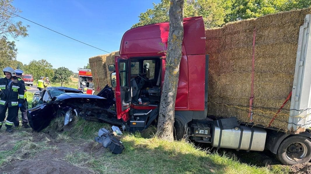 Rozbity samochód osobowy po zderzeniu z ciężarówką na poboczu drogi, na miejscu straż pożarna i ratownicy.