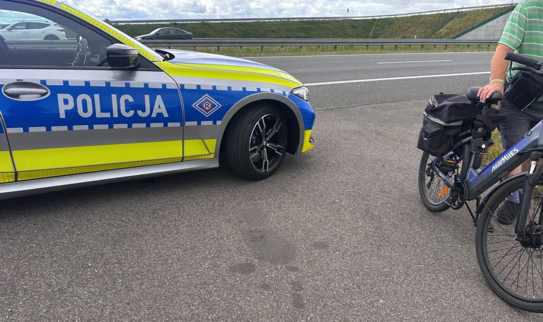 Policjant zatrzymuje rowerzystę na drodze ekspresowej S7, obok samochód policyjny i rower elektryczny.
