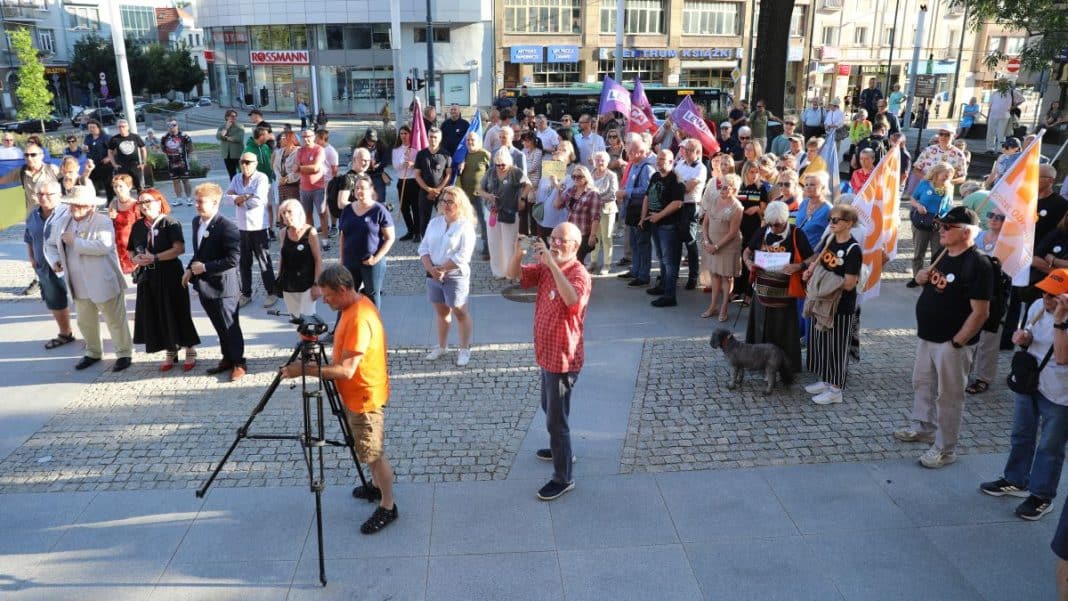 Uczestnicy zgromadzenia przed olsztyńskim ratuszem trzymają flagi Ukrainy i UE, niektórzy z transparentami, w słoneczny dzień.
