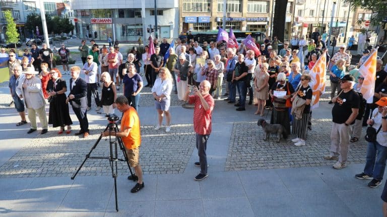 Uczestnicy zgromadzenia przed olsztyńskim ratuszem trzymają flagi Ukrainy i UE, niektórzy z transparentami, w słoneczny dzień.
